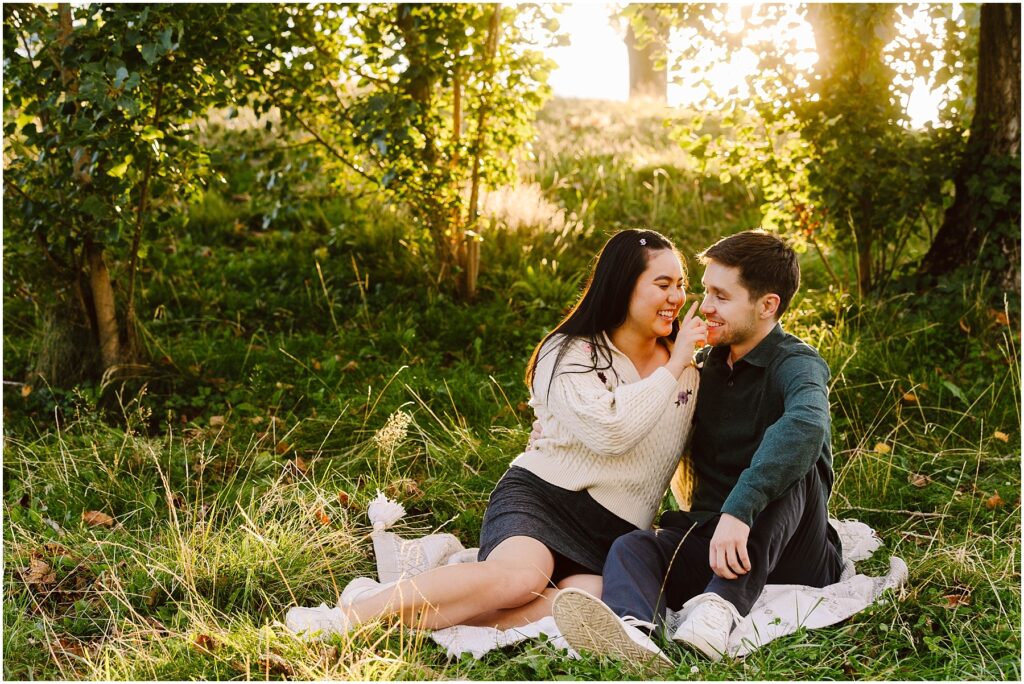 Snohomish Wedding Photographer GSquared Weddings A couple sits on a blanket in a sunlit forest clearing, smiling at each other with the joy of their newfound engagement. The woman wears a white sweater and the man a dark green shirt, surrounded by grass and trees as sunlight filters through the leaves. Serving Seattle, Snohomish and Washington State
