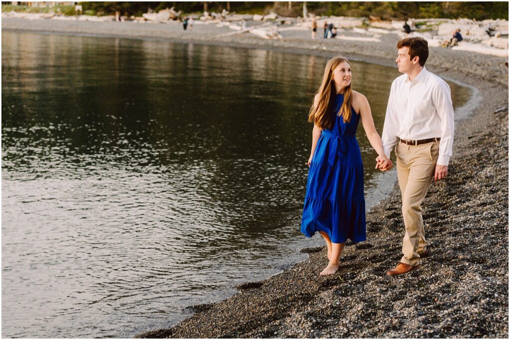 Snohomish Wedding Photographer GSquared Weddings A couple walks hand in hand along a pebbled beach. The woman wears a blue dress, and the man wears a white shirt and beige pants. The calm water reflects the evening light, and driftwood is scattered along the shore. Serving Seattle, Snohomish and Washington State