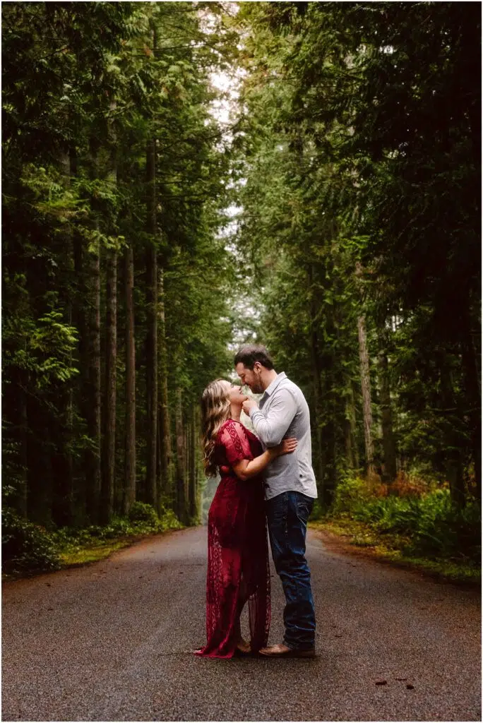Snohomish Wedding Photographer GSquared Weddings A couple embraces and kisses on a forest road surrounded by tall dense trees The woman in a deep red dress and the man in a light shirt and jeans share a tender engagement moment The serene scene is romantic with lush greenery creating a natural corridor Serving Seattle Snohomish and Washington State