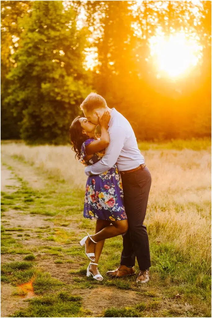 Snohomish Wedding Photographer GSquared Weddings A couple embraces in a sunlit field celebrating their engagement The woman in a floral dress and white sandals kisses her fiancé who is dressed in a light shirt and dark pants As the sun sets behind them it casts a warm golden light over this joyful moment Serving Seattle Snohomish and Washington State