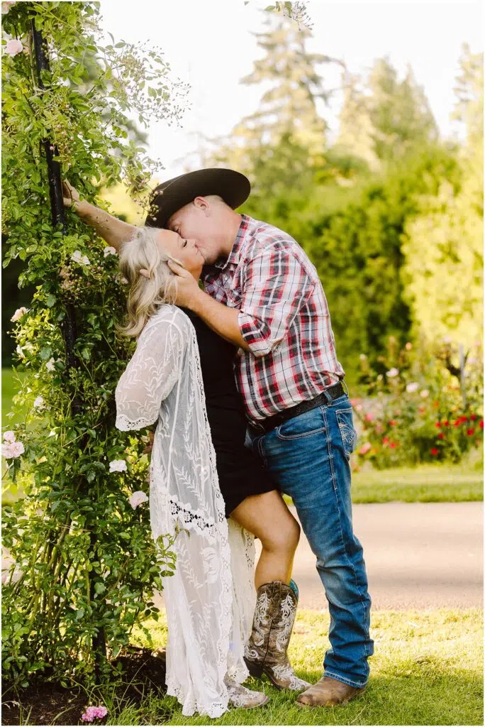 Snohomish Wedding Photographer GSquared Weddings A couple shares a romantic engagement kiss in a sunny garden The man sporting a cowboy hat and plaid shirt leans against an archway draped in greenery The woman in a white lace overlay and boots lovingly embraces him The lush background hints at the warmth of summertime Serving Seattle Snohomish and Washington State