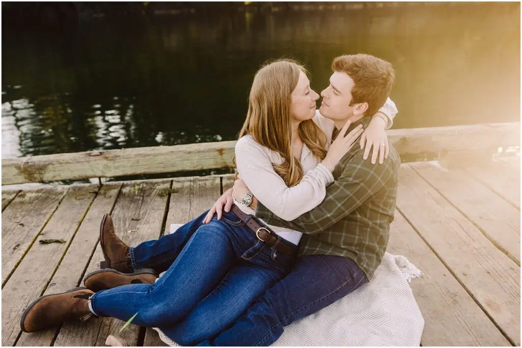 Snohomish Wedding Photographer GSquared Weddings A couple sits on a blanket on a wooden dock by the water their embrace signaling the joy of engagement They gaze at each other with the womans arms wrapped around the mans neck The setting is peaceful sunlight softly illuminating this special moment Serving Seattle Snohomish and Washington State