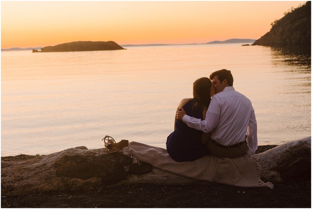 Snohomish Wedding Photographer GSquared Weddings A couple sits on a blanket at a beach during sunset, embracing as they look at the calm sea. A pair of sandals is placed nearby, and rocky islands are visible in the distance against the warm, glowing sky. Serving Seattle, Snohomish and Washington State
