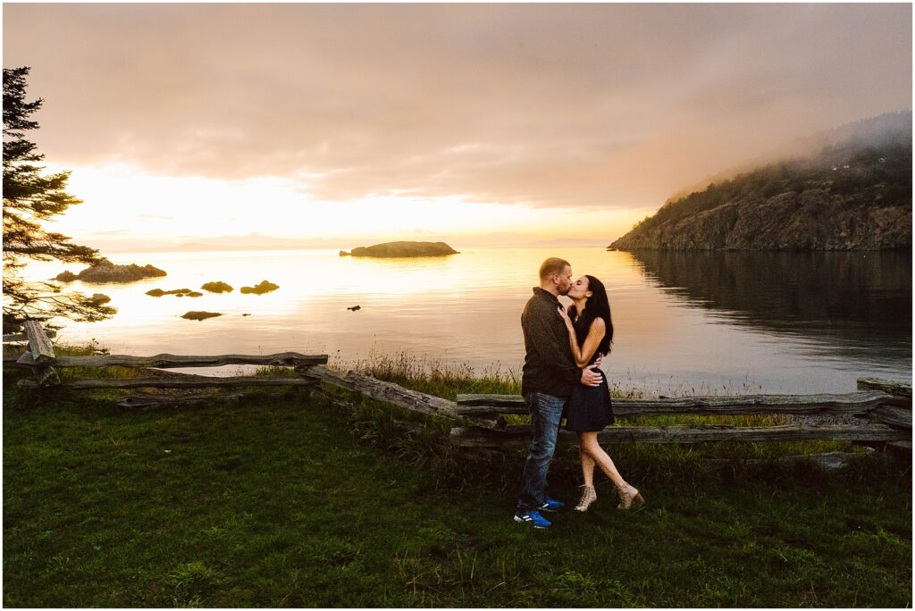 Snohomish Wedding Photographer GSquared Weddings A couple embraces and kisses by a wooden fence overlooking a serene lake at sunset. The sky is colorful, with clouds and a glowing horizon. Rocky hills and small islands are visible in the background, reflecting on the calm water. Serving Seattle, Snohomish and Washington State