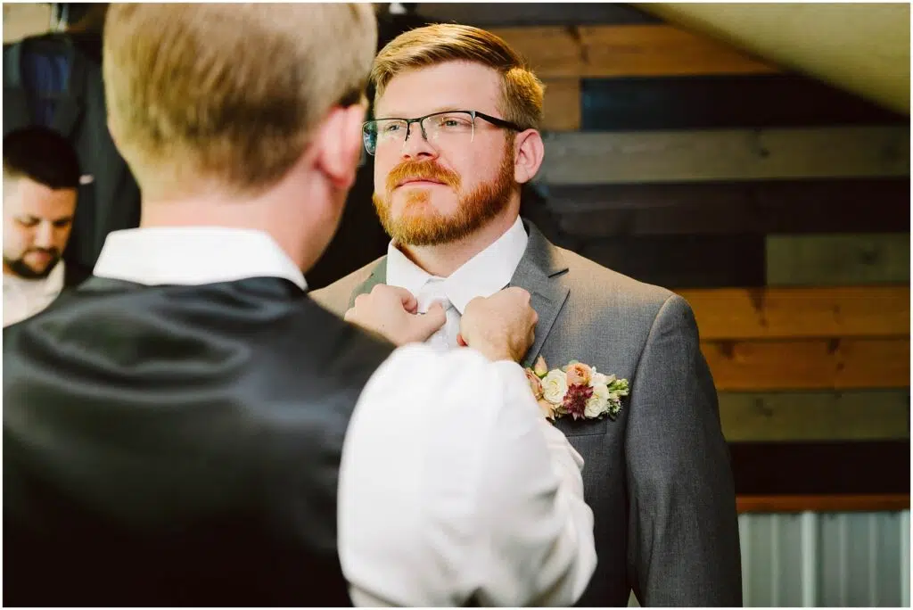 Snohomish Wedding Photographer GSquared Weddings A groom with a beard stands facing forward wearing a gray suit and a floral boutonniere Another person seen from behind adjusts the grooms bow tie In the background wood panels and another person are visible Serving Seattle Snohomish and Washington State Snohomish Wedding Photographer GSquared Weddings A groom with a beard stands facing forward, wearing a gray suit and a floral boutonniere. Another person, seen from behind, adjusts the grooms bow tie. In the background, wood panels and another person are visible. Serving Seattle, Snohomish and Washington State