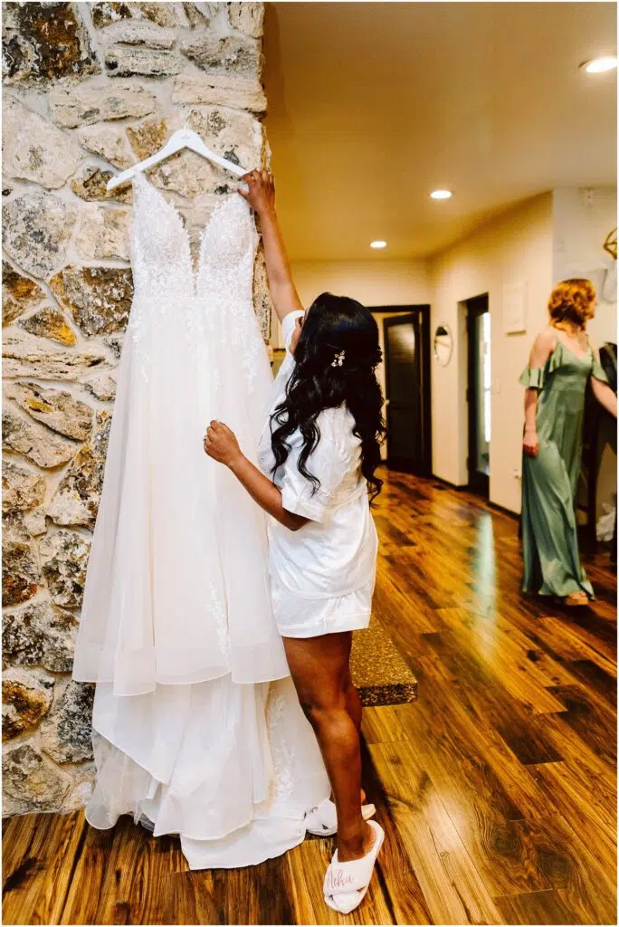 Snohomish Wedding Photographer GSquared Weddings A woman with long dark hair wearing a white robe and slippers reaches toward a white wedding dress hanging on a stone wall The room has wooden floors and another person is seen in the background wearing a green dress Serving Seattle Snohomish and Washington State Snohomish Wedding Photographer GSquared Weddings A woman with long dark hair, wearing a white robe and slippers, reaches toward a white wedding dress hanging on a stone wall. The room has wooden floors and another person is seen in the background wearing a green dress. Serving Seattle, Snohomish and Washington State