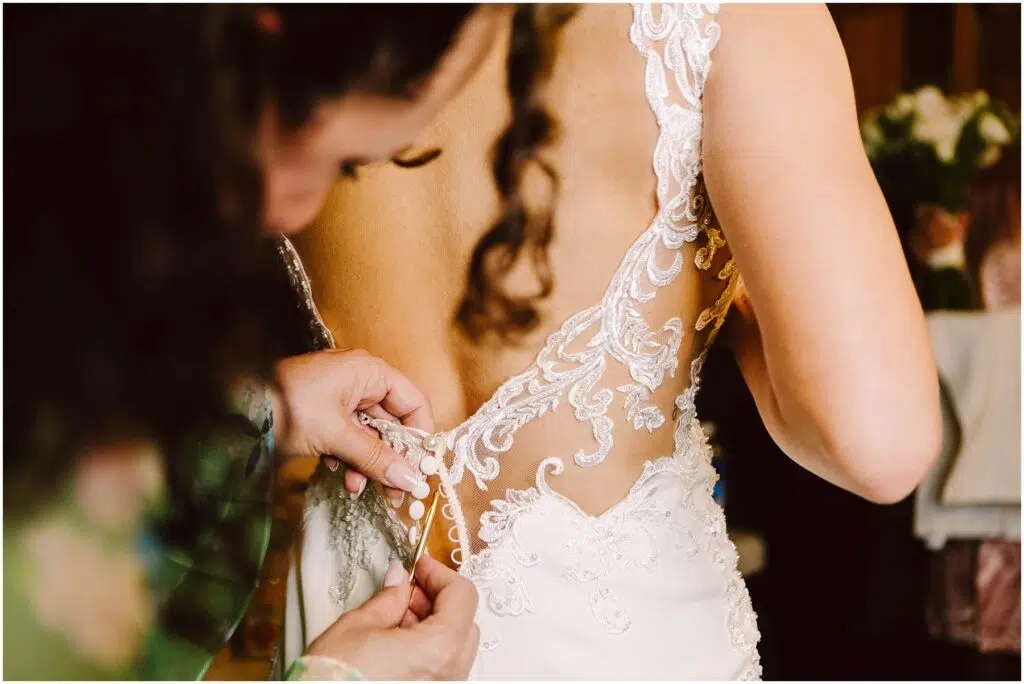 Snohomish Wedding Photographer GSquared Weddings A closeup of a bride in a lace wedding dress being buttoned up by an assistant Her back is facing the camera revealing intricate lace details on the dress Serving Seattle Snohomish and Washington State Snohomish Wedding Photographer GSquared Weddings A close-up of a bride in a lace wedding dress being buttoned up by an assistant. Her back is facing the camera, revealing intricate lace details on the dress. Serving Seattle, Snohomish and Washington State