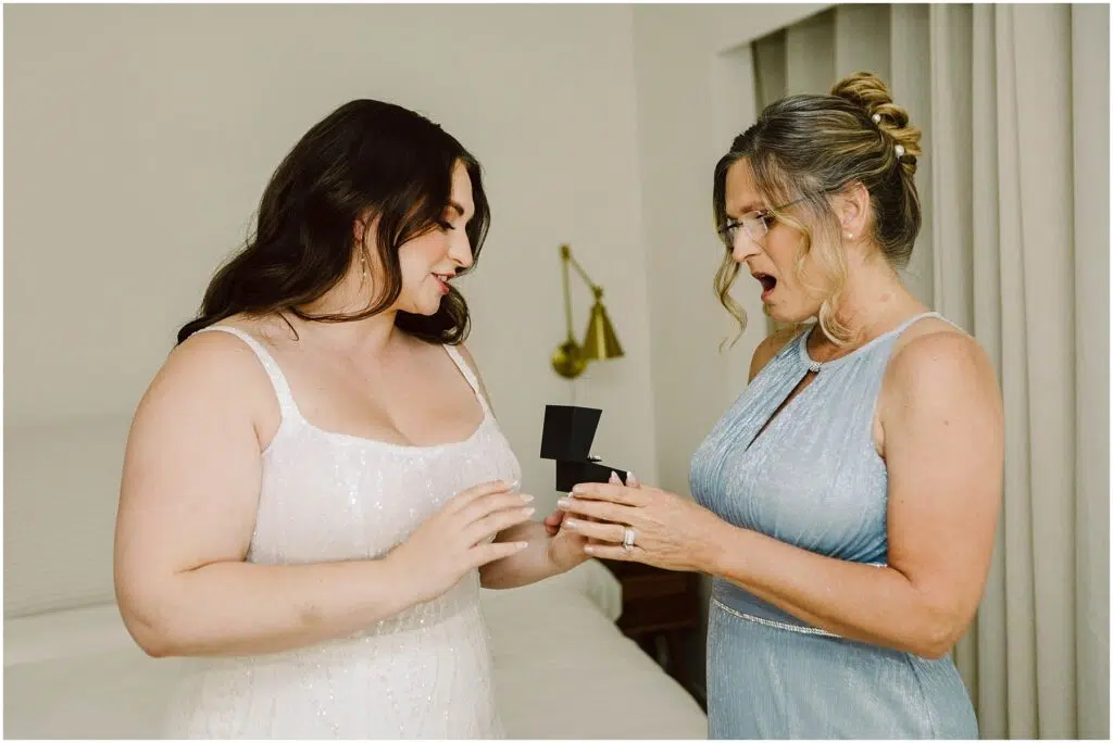 Snohomish Wedding Photographer GSquared Weddings Two women stand in a room one in a white dress holding an open black box the other in a light blue dress looks surprised and joyful They appear to be celebrating a special occasion A bed and a wall lamp are in the background Serving Seattle Snohomish and Washington State Snohomish Wedding Photographer GSquared Weddings Two women stand in a room, one in a white dress holding an open black box; the other in a light blue dress looks surprised and joyful. They appear to be celebrating a special occasion. A bed and a wall lamp are in the background. Serving Seattle, Snohomish and Washington State
