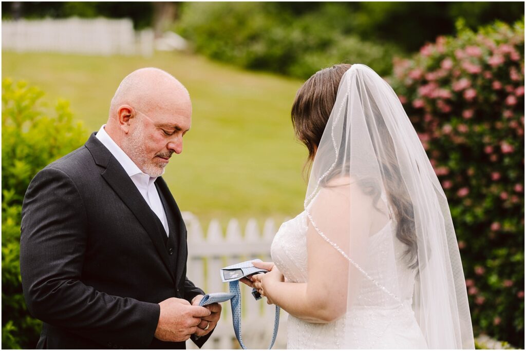 Bride in a veil gives a father figure a blue tie with a small photo patch before the ceremony Snohomish wedding photographer gsquared weddings a bride and a man in a suit possibly her father stand outdoors in a garden setting The bride facing away holds a piece of paper The man looks down at a tie in his hands A white fence and greenery are in the background Serving seattle snohomish and washington state