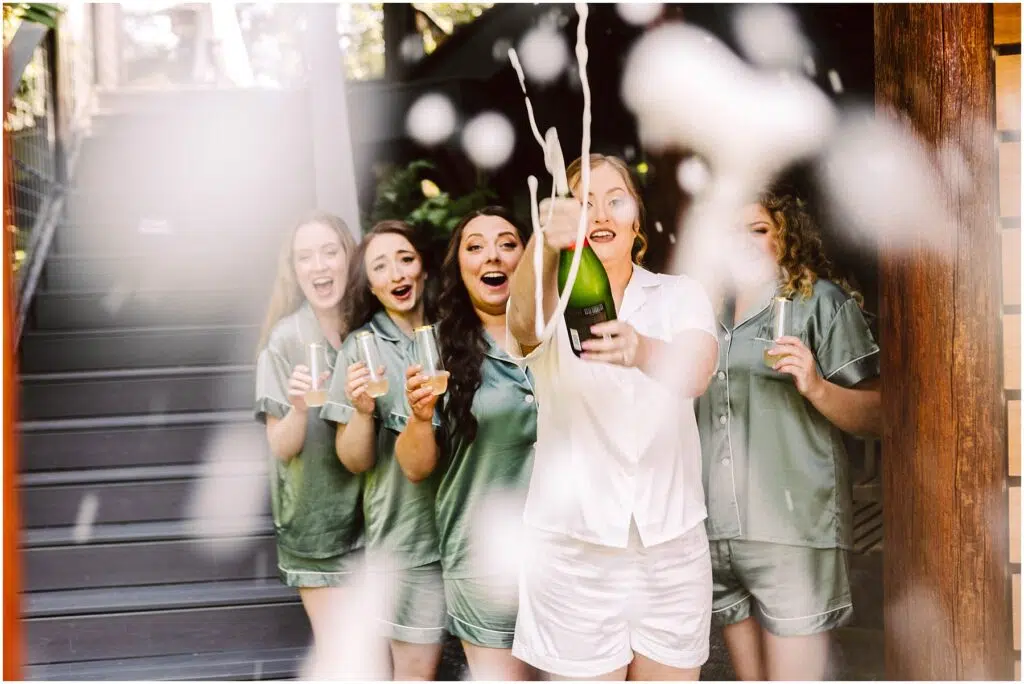 Snohomish Wedding Photographer GSquared Weddings A group of five women in matching pajamas one in white and the others in green are excitedly popping a bottle of champagne outdoors The cork flies and champagne sprays into the air capturing a moment of joyful celebration Serving Seattle Snohomish and Washington State Snohomish Wedding Photographer GSquared Weddings A group of five women in matching pajamas, one in white and the others in green, are excitedly popping a bottle of champagne outdoors. The cork flies and champagne sprays into the air, capturing a moment of joyful celebration. Serving Seattle, Snohomish and Washington State