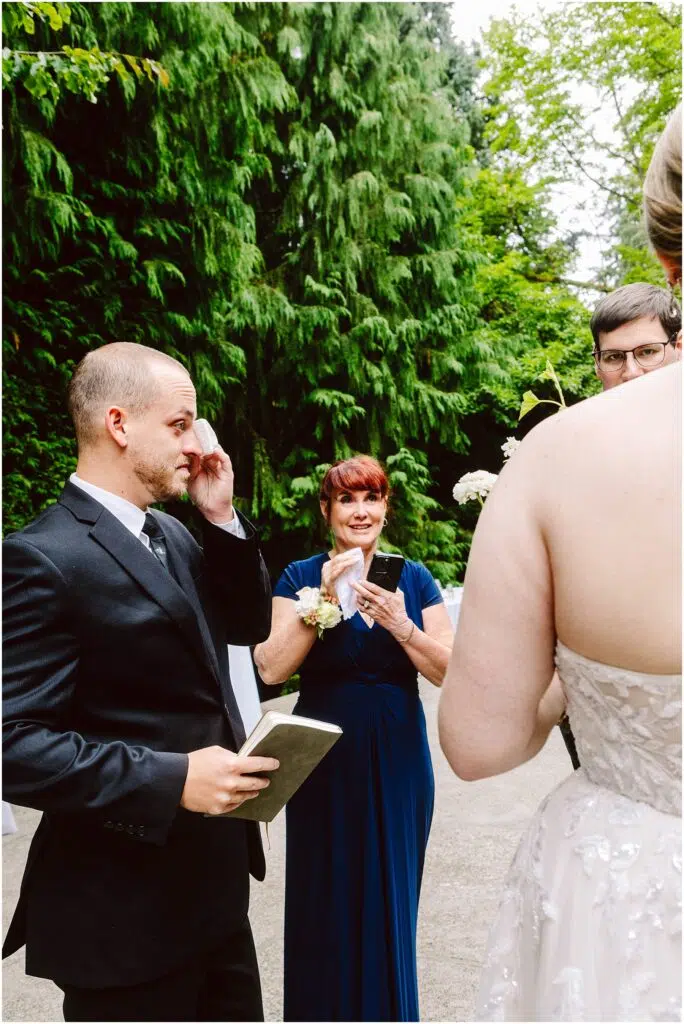 Snohomish Wedding Photographer GSquared Weddings A groom in a suit stands outdoors holding a book and wiping a tear A woman in a blue dress smiling holds a smartphone The bride in a white dress is partially visible Lush green foliage surrounds them Serving Seattle Snohomish and Washington State Snohomish Wedding Photographer GSquared Weddings A groom in a suit stands outdoors, holding a book and wiping a tear. A woman in a blue dress, smiling, holds a smartphone. The bride, in a white dress, is partially visible. Lush green foliage surrounds them. Serving Seattle, Snohomish and Washington State