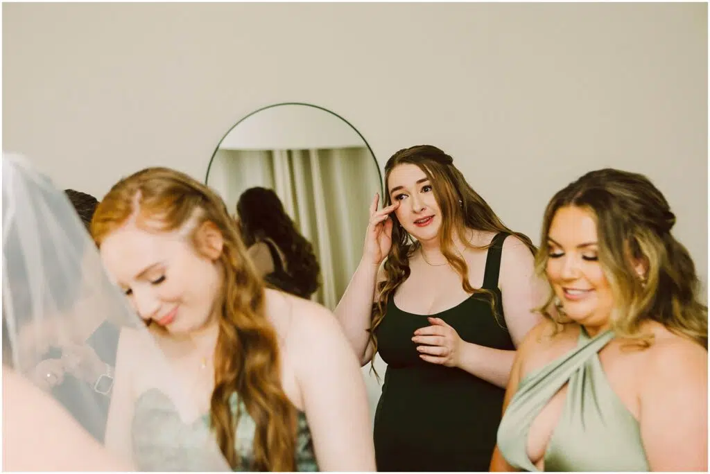 Snohomish Wedding Photographer GSquared Weddings Three women in elegant dresses are in a softly lit room two of them smiling and looking down while one wipes her eye gently A round mirror is in the background reflecting part of the scene Serving Seattle Snohomish and Washington State Snohomish Wedding Photographer GSquared Weddings Three women in elegant dresses are in a softly lit room, two of them smiling and looking down, while one wipes her eye gently. A round mirror is in the background, reflecting part of the scene. Serving Seattle, Snohomish and Washington State