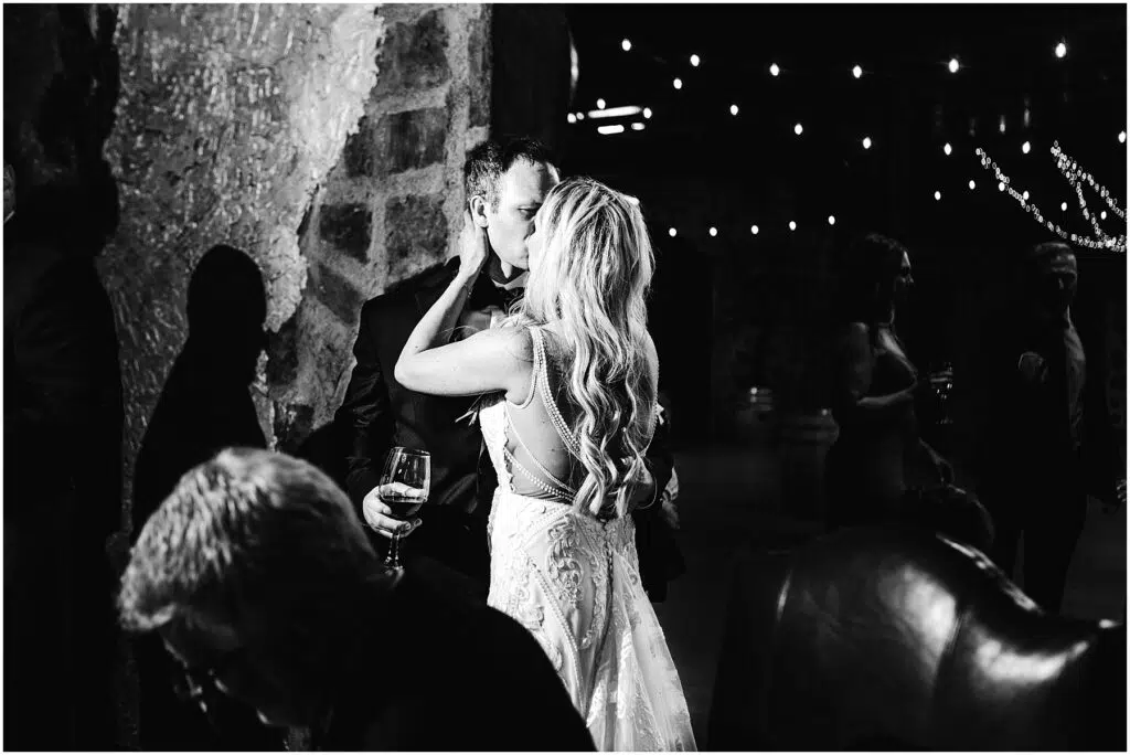 Snohomish Wedding Photographer GSquared Weddings A couple in formal attire shares a kiss at an indoor event The scene is lit by string lights creating a romantic atmosphere Other people are visible in the background and a seated man is in the foreground Black and white photo Serving Seattle Snohomish and Washington State Snohomish Wedding Photographer GSquared Weddings A couple in formal attire shares a kiss at an indoor event. The scene is lit by string lights, creating a romantic atmosphere. Other people are visible in the background, and a seated man is in the foreground. Black and white photo. Serving Seattle, Snohomish and Washington State