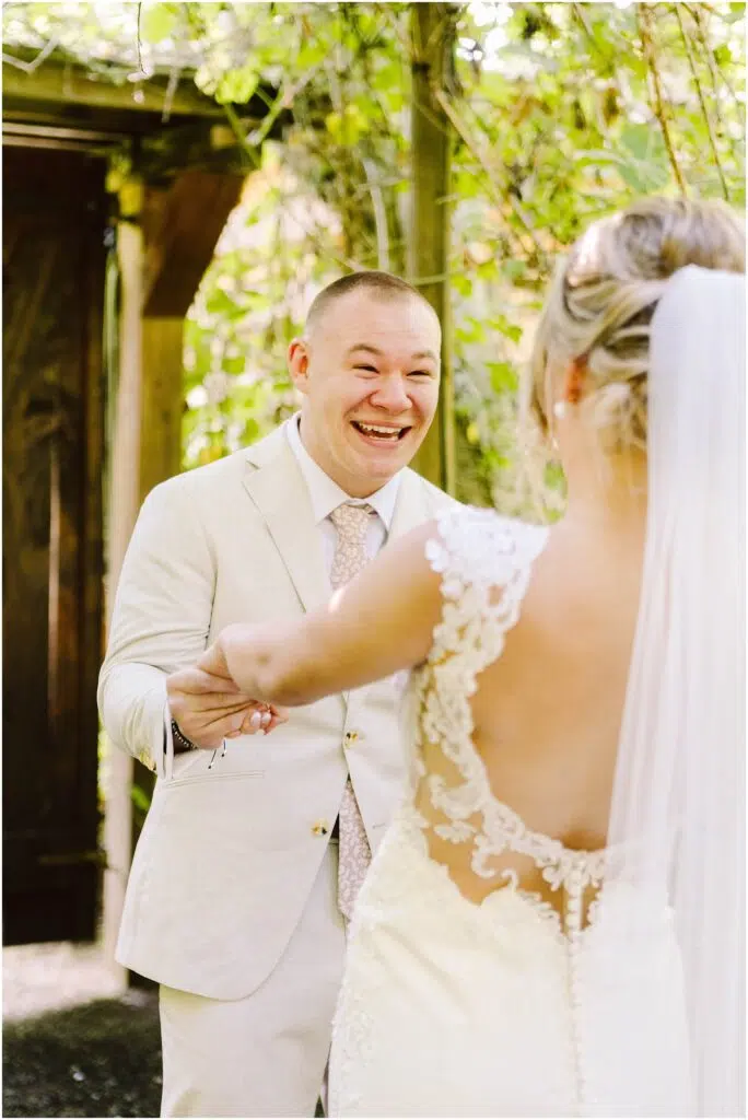 Snohomish Wedding Photographer GSquared Weddings Groom in a light beige suit smiling joyfully holding hands with a bride in a lace dress with a veil facing him They are outdoors surrounded by green foliage and wooden structures Serving Seattle Snohomish and Washington State Snohomish Wedding Photographer GSquared Weddings Groom in a light beige suit smiling joyfully, holding hands with a bride in a lace dress with a veil, facing him. They are outdoors, surrounded by green foliage and wooden structures. Serving Seattle, Snohomish and Washington State