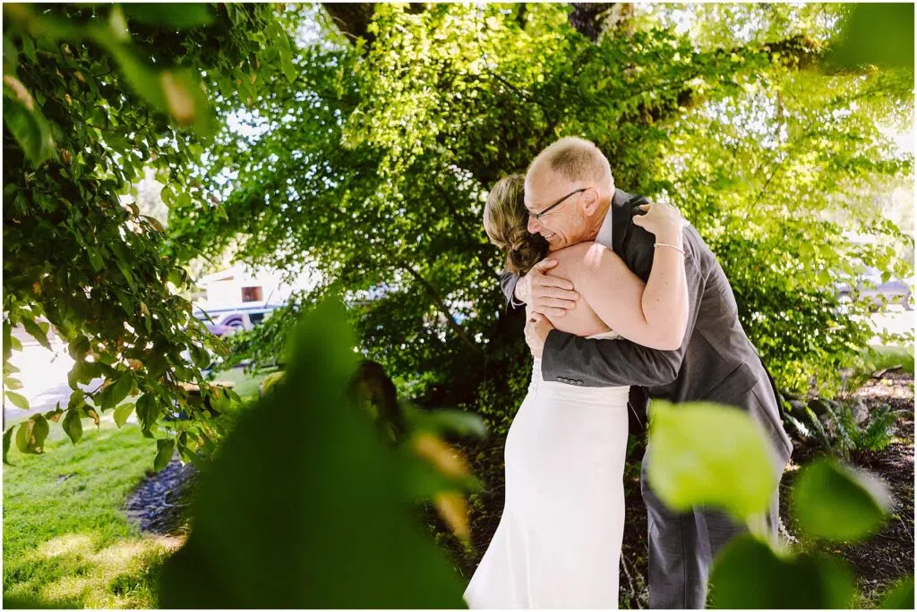 Snohomish Wedding Photographer GSquared Weddings A bride in a white dress hugs an older man in a suit amidst lush greenery Sunlight filters through the leaves casting soft light on the scene The foliage frames the couple creating an intimate joyful moment Serving Seattle Snohomish and Washington State Snohomish Wedding Photographer GSquared Weddings A bride in a white dress hugs an older man in a suit amidst lush greenery. Sunlight filters through the leaves, casting soft light on the scene. The foliage frames the couple, creating an intimate, joyful moment. Serving Seattle, Snohomish and Washington State