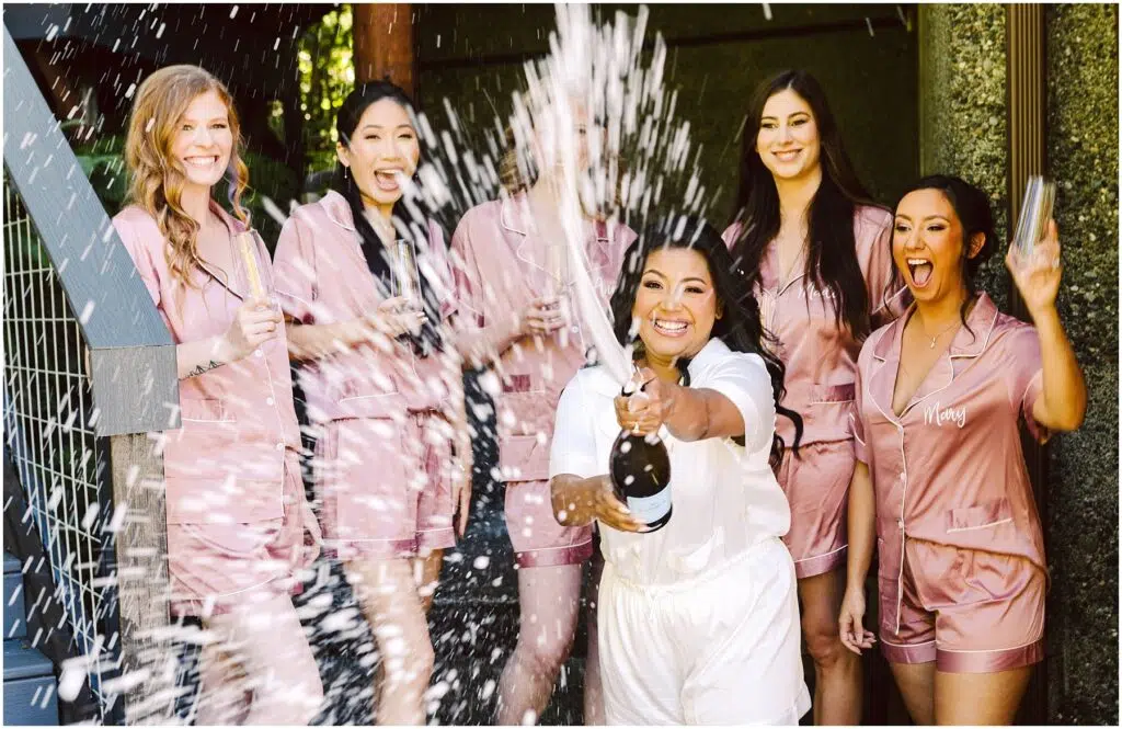 Snohomish Wedding Photographer GSquared Weddings A woman in white pajamas joyfully pops open a champagne bottle spraying it around her Five women in matching pink pajamas laugh and react with excitement They are outside with greenery and a wooden structure in the background Serving Seattle Snohomish and Washington State Snohomish Wedding Photographer GSquared Weddings A woman in white pajamas joyfully pops open a champagne bottle, spraying it around her. Five women in matching pink pajamas laugh and react with excitement. They are outside, with greenery and a wooden structure in the background. Serving Seattle, Snohomish and Washington State
