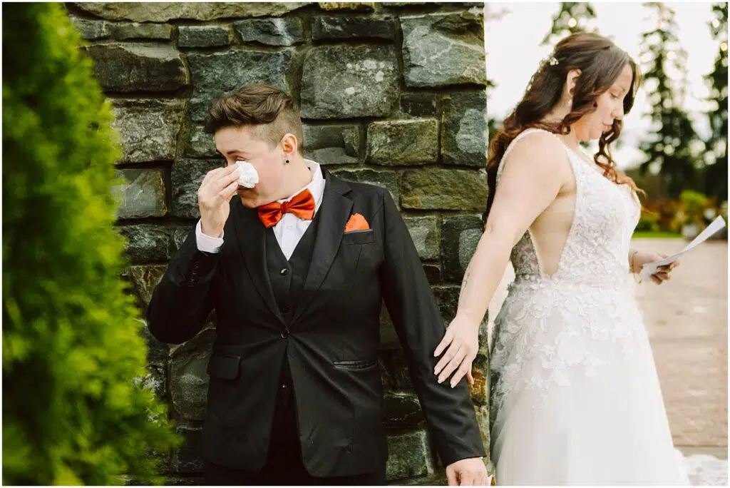 Snohomish Wedding Photographer GSquared Weddings A couple stands near a stone wall both in formal attire The person in a suit with a red bow tie wipes tears with a tissue The other in a white lace gown gently touches their partners hand holding a paper Emotional moment in a wedding setting Serving Seattle Snohomish and Washington State Snohomish Wedding Photographer GSquared Weddings A couple stands near a stone wall, both in formal attire. The person in a suit with a red bow tie wipes tears with a tissue. The other, in a white lace gown, gently touches their partners hand, holding a paper. Emotional moment in a wedding setting. Serving Seattle, Snohomish and Washington State