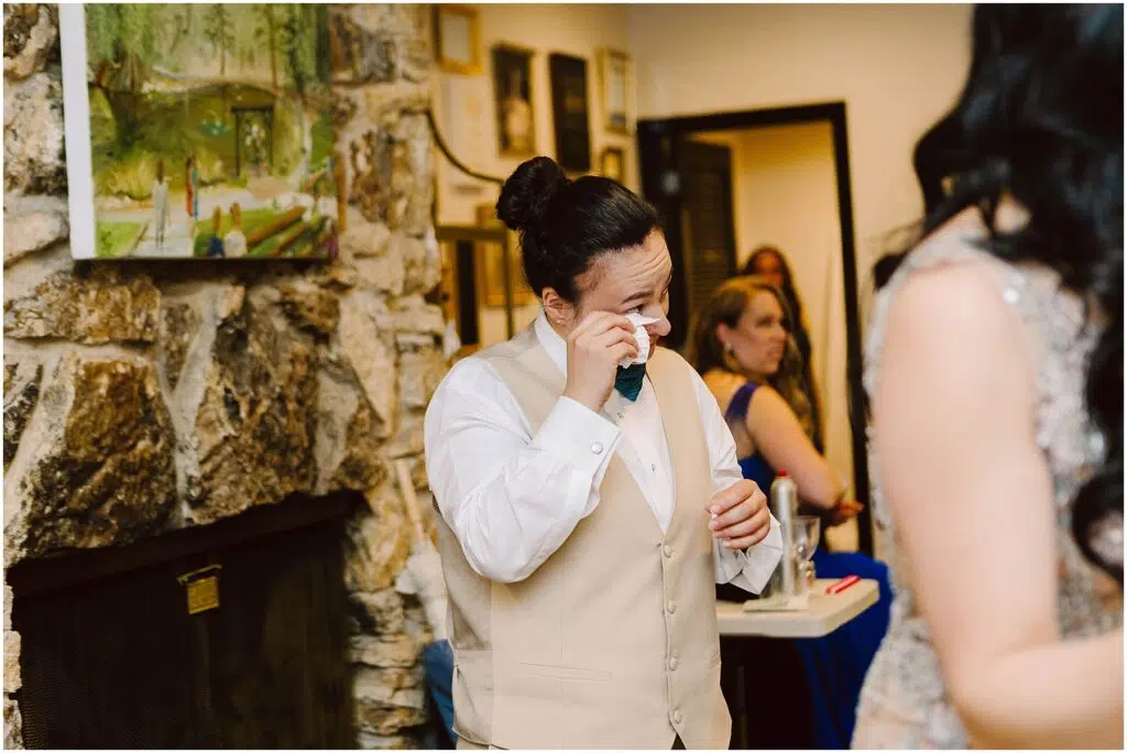 Snohomish Wedding Photographer GSquared Weddings A person in formal attire wipes away tears while standing near a stone fireplace They wear a cream vest and a blue bow tie A person in blue is seated in the background and a framed painting hangs on the wall Serving Seattle Snohomish and Washington State Snohomish Wedding Photographer GSquared Weddings A person in formal attire wipes away tears while standing near a stone fireplace. They wear a cream vest and a blue bow tie. A person in blue is seated in the background, and a framed painting hangs on the wall. Serving Seattle, Snohomish and Washington State
