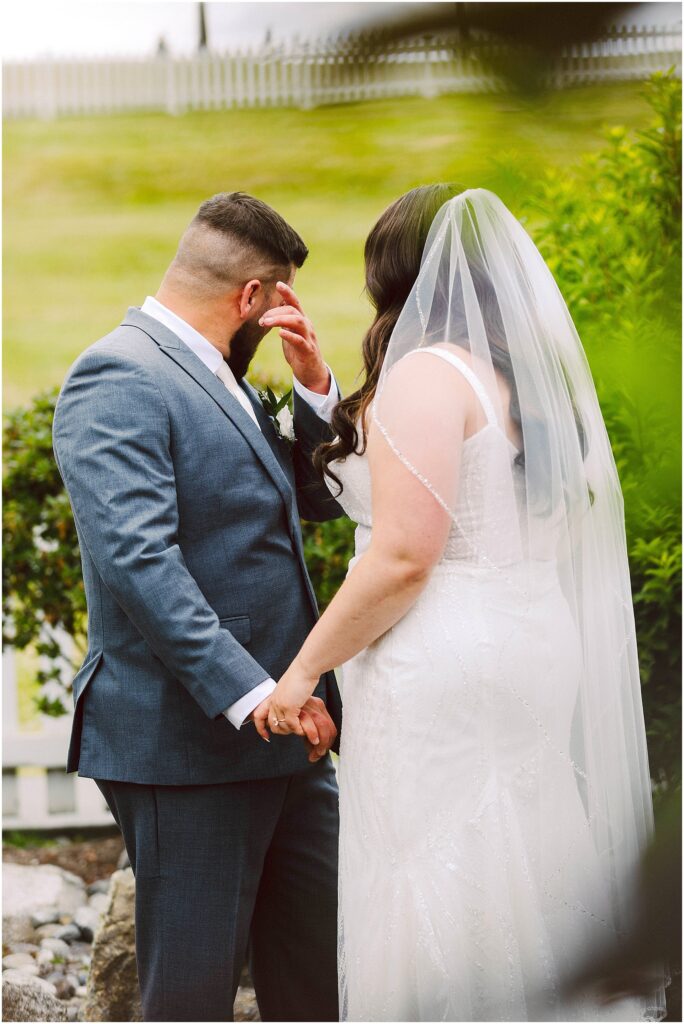 Groom wipes away tears while holding hands with the bride in her veil and gown outdoors Groom wipes away tears while holding hands with the bride in her veil and gown outdoors