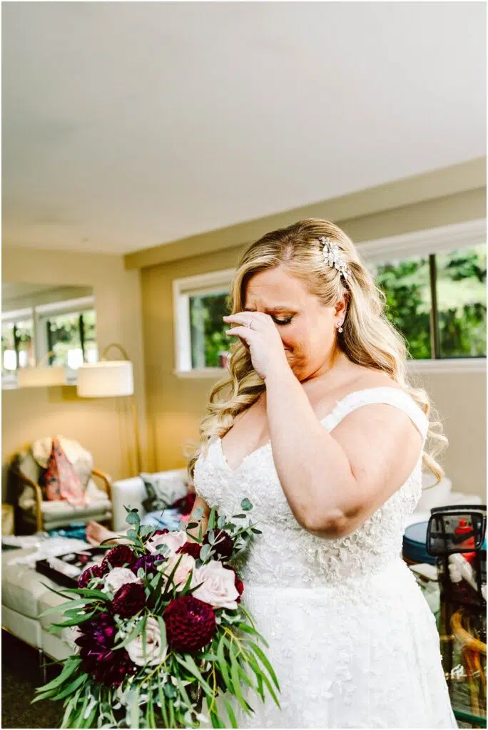 Snohomish Wedding Photographer GSquared Weddings A bride in a white lace gown and floral hairpiece wipes tears from her eyes holding a bouquet with dark red and white flowers She stands in a brightly lit room with modern furniture and large windows Serving Seattle Snohomish and Washington State Snohomish Wedding Photographer GSquared Weddings A bride in a white lace gown and floral hairpiece wipes tears from her eyes, holding a bouquet with dark red and white flowers. She stands in a brightly lit room with modern furniture and large windows. Serving Seattle, Snohomish and Washington State