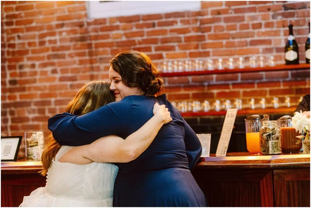 Snohomish Wedding Photographer GSquared Weddings Two women embrace warmly in front of a brick wall One wears a white dress the other a blue dress Behind them is a wooden counter with glassware and drink dispensers suggesting a celebratory event or gathering Serving Seattle Snohomish and Washington State Snohomish Wedding Photographer GSquared Weddings Two women embrace warmly in front of a brick wall. One wears a white dress, the other a blue dress. Behind them is a wooden counter with glassware and drink dispensers, suggesting a celebratory event or gathering. Serving Seattle, Snohomish and Washington State