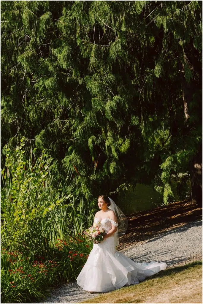 Snohomish Wedding Photographer GSquared Weddings A bride in a white wedding dress and veil walks on a gravel path holding a bouquet of flowers She is surrounded by lush green trees and plants with sunlight filtering through the leaves creating a serene outdoor setting Serving Seattle Snohomish and Washington State Snohomish Wedding Photographer GSquared Weddings A bride in a white wedding dress and veil walks on a gravel path, holding a bouquet of flowers. She is surrounded by lush green trees and plants, with sunlight filtering through the leaves, creating a serene outdoor setting. Serving Seattle, Snohomish and Washington State