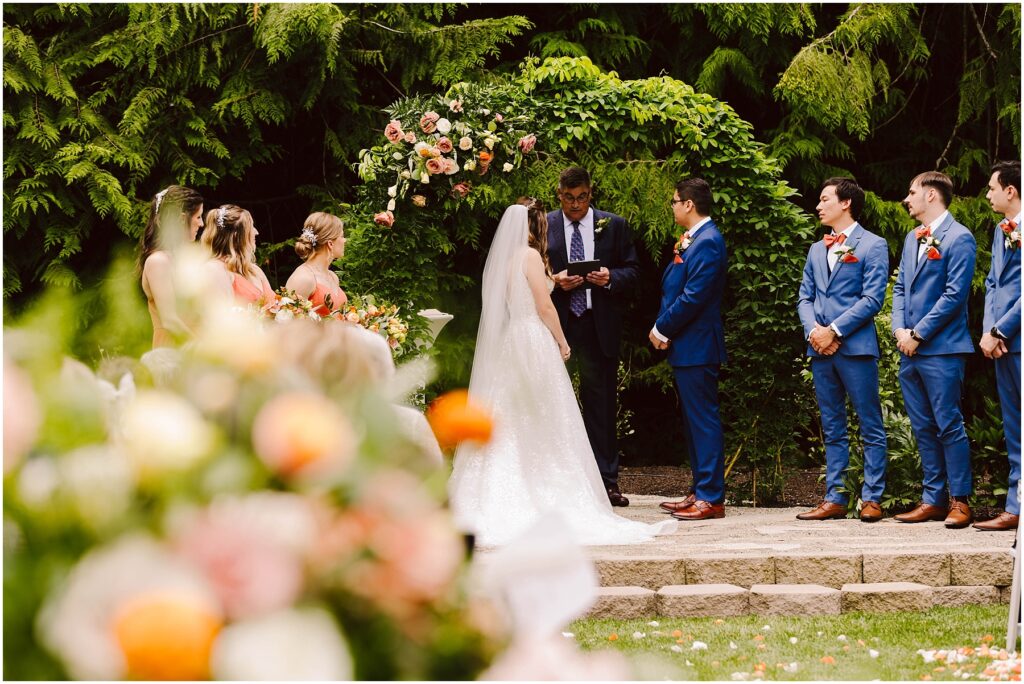 Snohomish Wedding Photographer GSquared Weddings Bride and groom stand facing each other under a floral arch during an outdoor wedding ceremony The officiant is reading while bridesmaids in pink and groomsmen in blue suits watch Floral arrangements and greenery surround the scene Serving Seattle Snohomish and Washington State Snohomish Wedding Photographer GSquared Weddings Bride and groom stand facing each other under a floral arch during an outdoor wedding ceremony. The officiant is reading, while bridesmaids in pink and groomsmen in blue suits watch. Floral arrangements and greenery surround the scene. Serving Seattle, Snohomish and Washington State