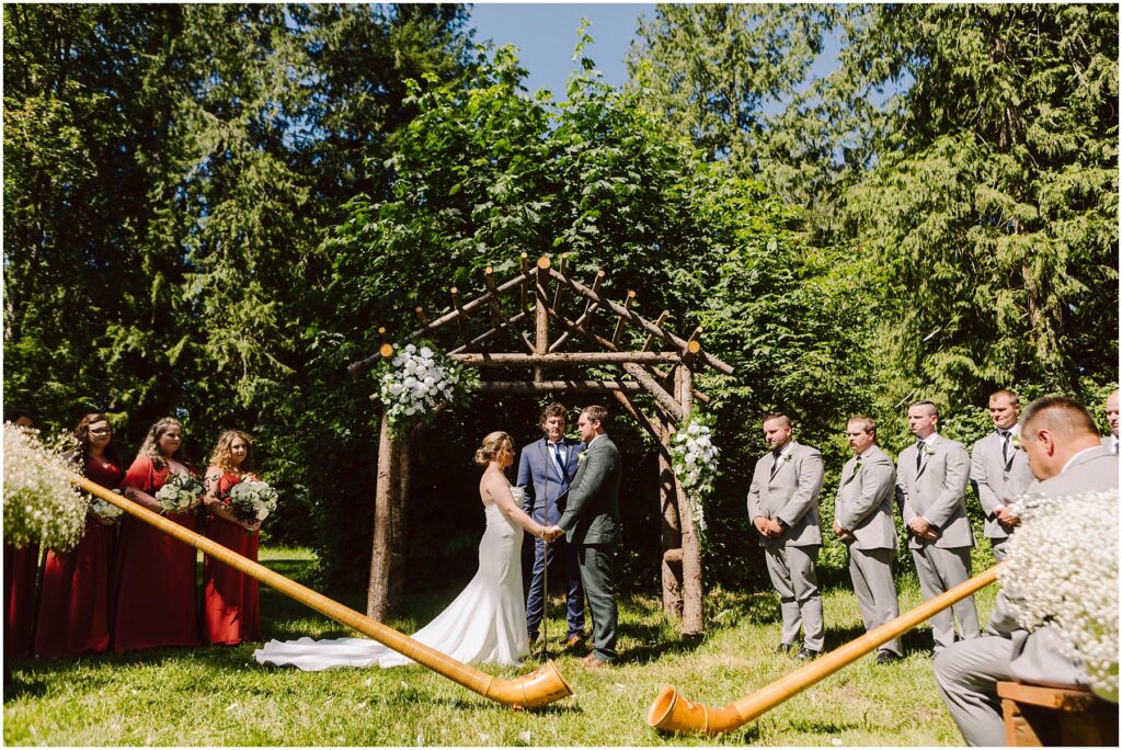 Snohomish Wedding Photographer GSquared Weddings A couple stands at a rustic wooden altar adorned with flowers in an outdoor setting surrounded by trees Bridesmaids in red dresses and groomsmen in gray suits stand on either side Two musicians play large wooden horns Serving Seattle Snohomish and Washington State Snohomish Wedding Photographer GSquared Weddings A couple stands at a rustic wooden altar adorned with flowers in an outdoor setting surrounded by trees. Bridesmaids in red dresses and groomsmen in gray suits stand on either side. Two musicians play large wooden horns. Serving Seattle, Snohomish and Washington State