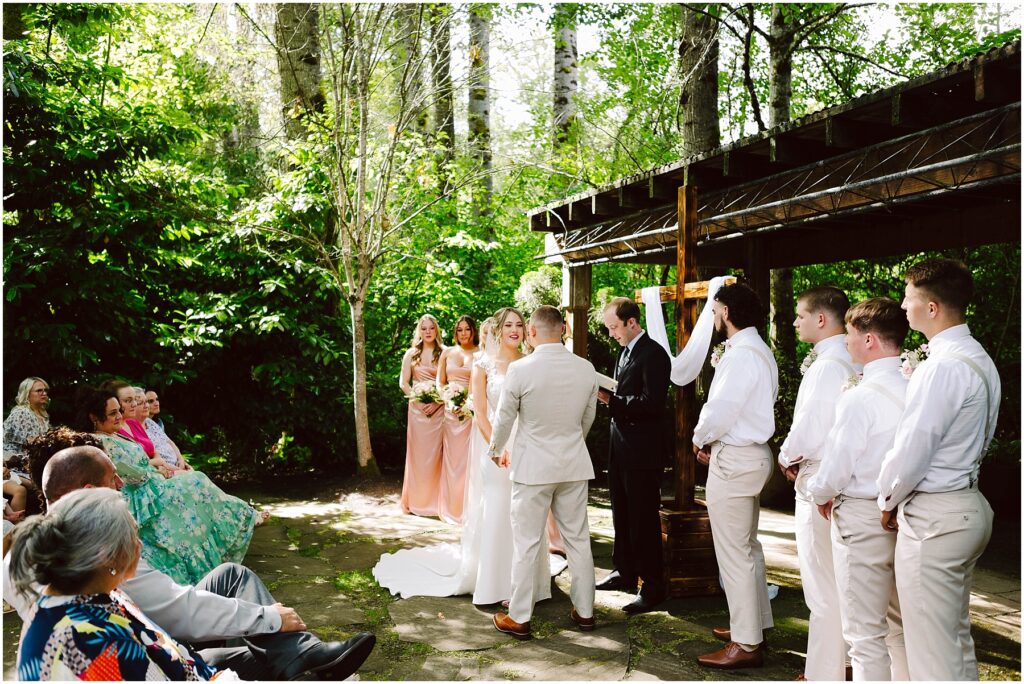 Snohomish Wedding Photographer GSquared Weddings An outdoor wedding ceremony in a lush green setting The bride and groom stand under a wooden pergola with bridesmaids in peach dresses and groomsmen in white shirts and khakis nearby Guests are seated watching attentively Serving Seattle Snohomish and Washington State Snohomish Wedding Photographer GSquared Weddings An outdoor wedding ceremony in a lush, green setting. The bride and groom stand under a wooden pergola, with bridesmaids in peach dresses and groomsmen in white shirts and khakis nearby. Guests are seated, watching attentively. Serving Seattle, Snohomish and Washington State
