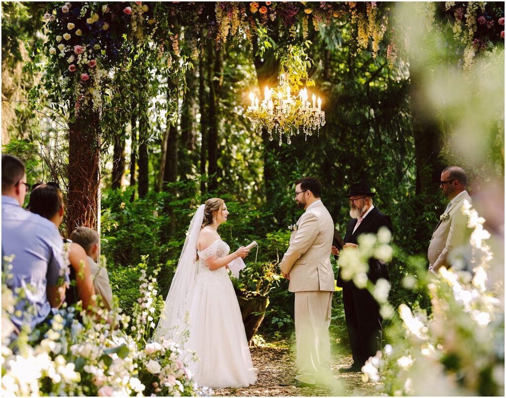 Snohomish Wedding Photographer GSquared Weddings A bride and groom stand under a floral arch exchanging vows in a lush green outdoor setting A chandelier hangs above them Guests and officiants are present with sunlight filtering through the trees Serving Seattle Snohomish and Washington State Snohomish Wedding Photographer GSquared Weddings A bride and groom stand under a floral arch, exchanging vows in a lush, green outdoor setting. A chandelier hangs above them. Guests and officiants are present, with sunlight filtering through the trees. Serving Seattle, Snohomish and Washington State