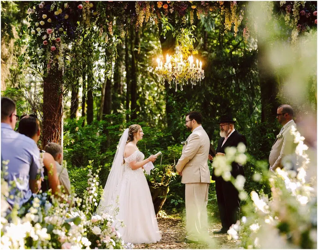 Snohomish Wedding Photographer GSquared Weddings A bride and groom stand under a floral arch exchanging vows in a lush green outdoor setting A chandelier hangs above them Guests and officiants are present with sunlight filtering through the trees Serving Seattle Snohomish and Washington State Snohomish Wedding Photographer GSquared Weddings A bride and groom stand under a floral arch, exchanging vows in a lush, green outdoor setting. A chandelier hangs above them. Guests and officiants are present, with sunlight filtering through the trees. Serving Seattle, Snohomish and Washington State