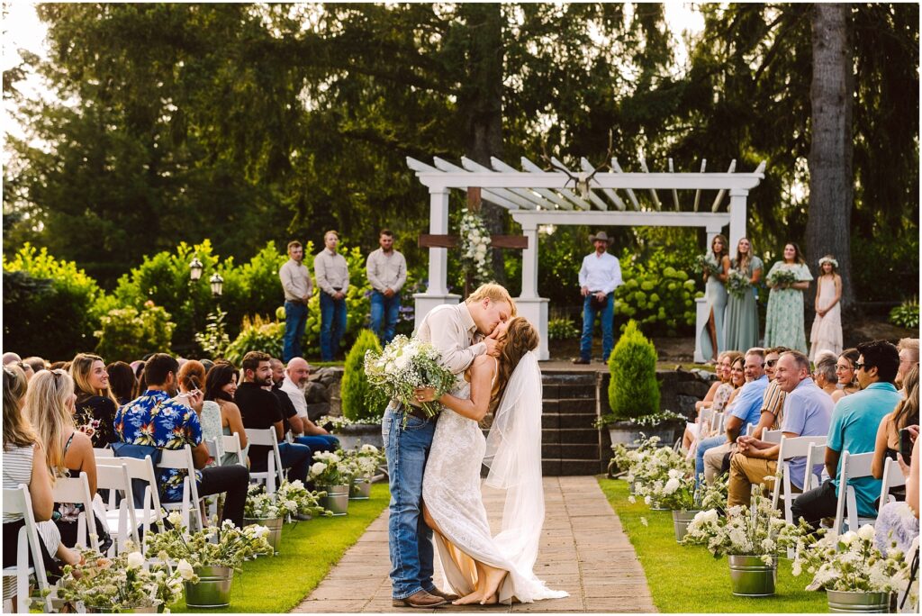 Snohomish Wedding Photographer GSquared Weddings A bride and groom share a kiss at an outdoor wedding ceremony The groom wears a light shirt and jeans while the bride is in a lace gown with a flowing veil Guests are seated along both sides of the aisle lined with flowers under a sunny sky Serving Seattle Snohomish and Washington State Snohomish Wedding Photographer GSquared Weddings A bride and groom share a kiss at an outdoor wedding ceremony. The groom wears a light shirt and jeans, while the bride is in a lace gown with a flowing veil. Guests are seated along both sides of the aisle, lined with flowers, under a sunny sky. Serving Seattle, Snohomish and Washington State