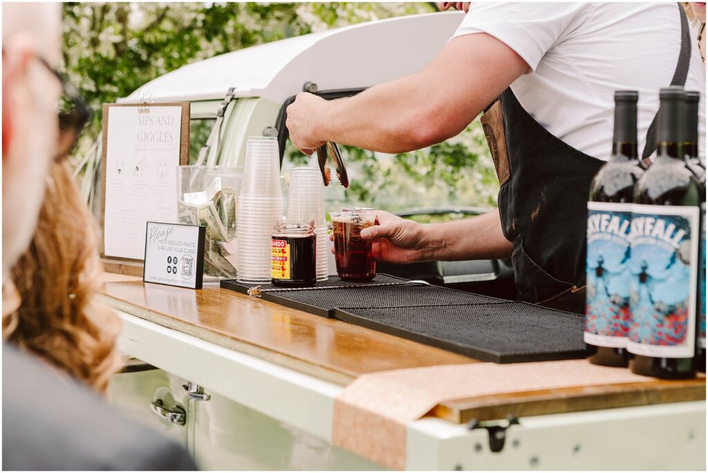 Snohomish wedding photographer gsquared weddings a bartender prepares drinks at an outdoor bar set up in a vintage vehicle The counter has cups and two bottles of wine with artistic labels A small sign and menu are nearby The blurred background shows greenery and a person watching Serving seattle snohomish and washington state