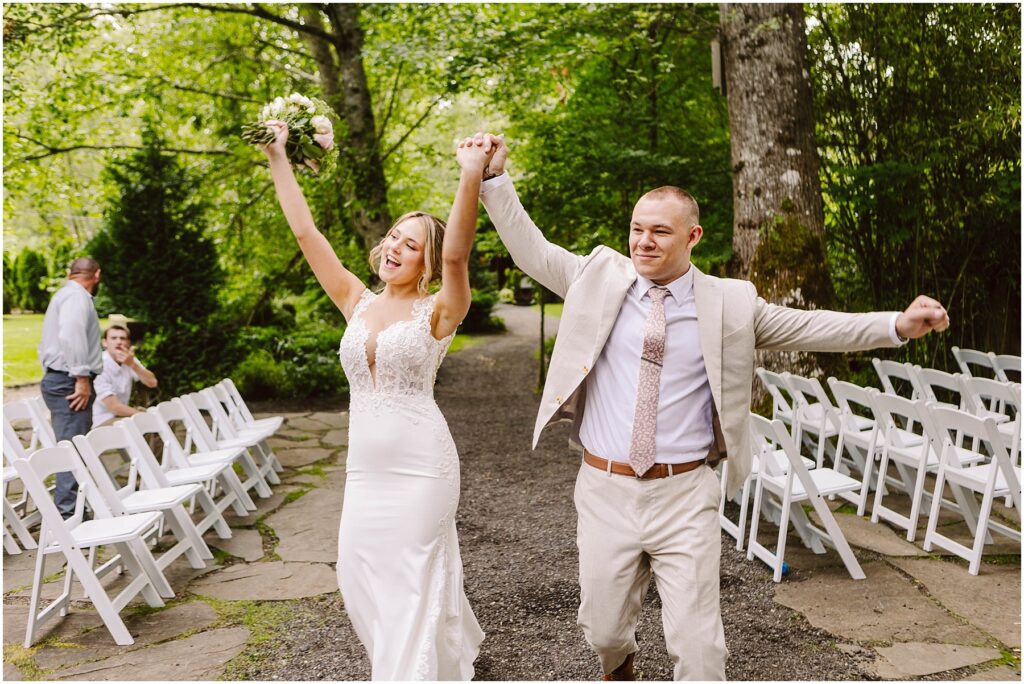 Snohomish wedding photographer gsquared weddings a bride in a lace wedding dress and a groom in a light suit joyfully walk down an outdoor aisle holding hands with arms raised Empty white chairs line the path Lush green trees surround the scene Serving seattle snohomish and washington state