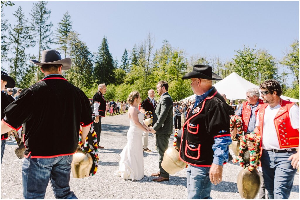 Snohomish wedding photographer gsquared weddings a bride and groom stand outdoors surrounded by men in traditional attire some wearing cowboy hats and holding large decorated cowbells Trees and a tent are in the background with clear blue skies above Serving seattle snohomish and washington state