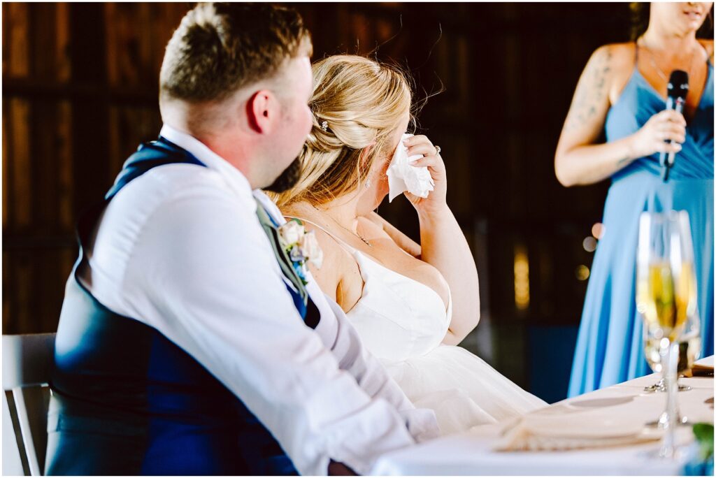 Snohomish wedding photographer gsquared weddings a bride wipes her tears with a tissue while sitting next to the groom at a wedding reception They are listening to a speech being given by a woman in a blue dress holding a microphone Champagne glasses are on the table Serving seattle snohomish and washington state