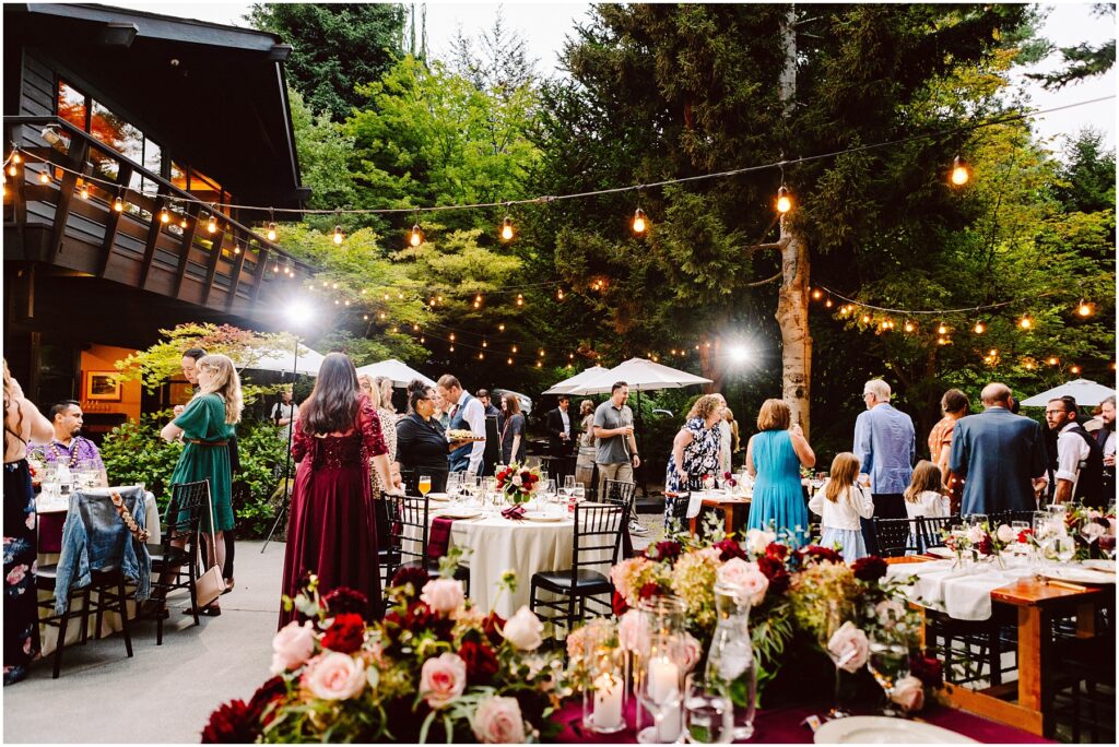 Snohomish wedding photographer gsquared weddings guests gather at an outdoor event with string lights and tables decorated with flowers People are engaged in conversation near a building and trees creating a warm festive atmosphere under a clear sky Serving seattle snohomish and washington state