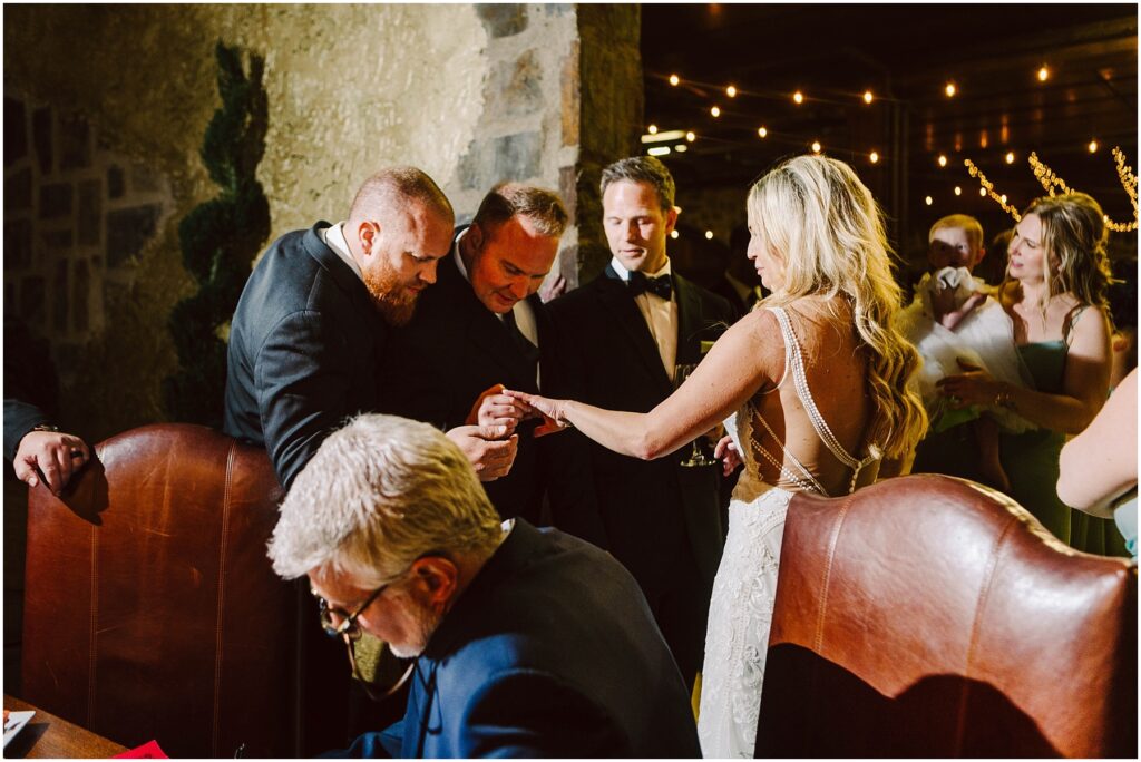 Snohomish wedding photographer gsquared weddings a bride in a white gown shows her ring to three men in suits at a dimly lit event One man leans in for a closer look while another holds a drink A seated older man in the foreground writes on paper String lights hang in the background Serving seattle snohomish and washington state