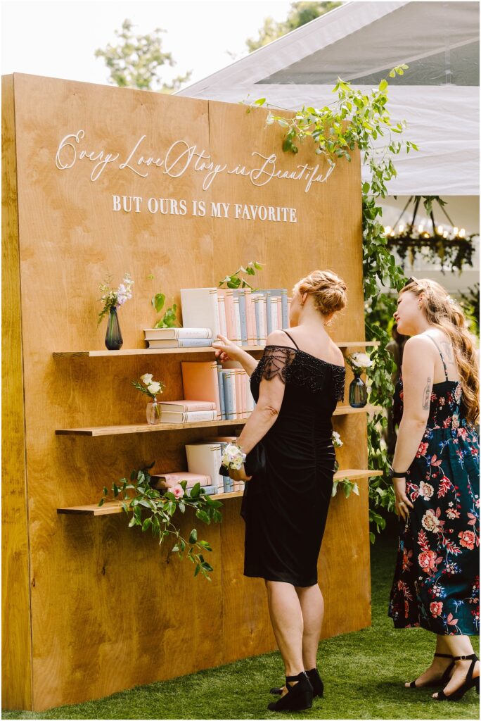 Snohomish wedding photographer gsquared weddings two women stand in front of a wooden bookshelf at an outdoor event The bookshelf is adorned with books and plants and a sign above reads every love story is beautiful but ours is my favorite The women are dressed in formal attire Serving seattle snohomish and washington state