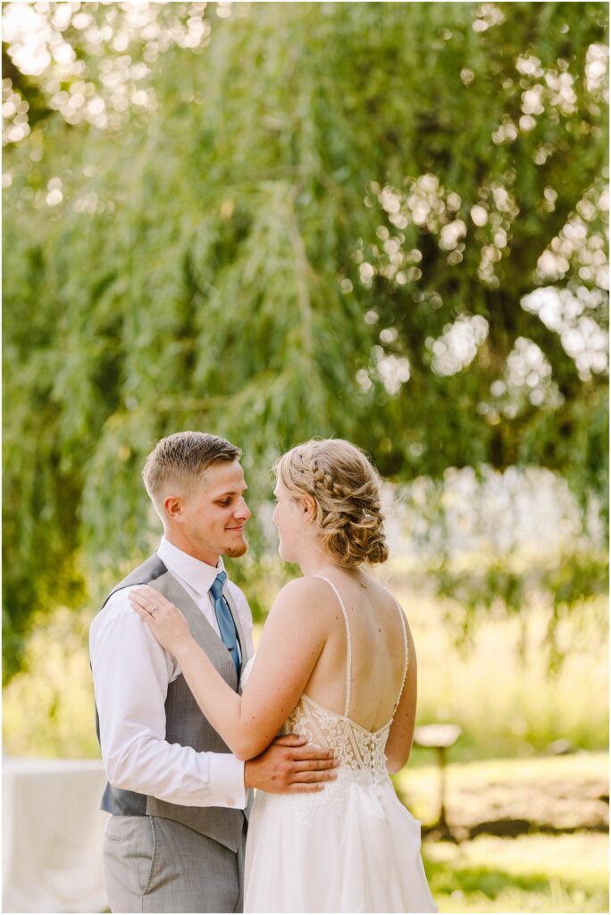 Snohomish wedding photographer gsquared weddings a couple dances outdoors beneath a large tree The woman wears a white dress with lace details and the man wears a gray vest with a blue tie Sunlight filters through the leaves creating a warm romantic atmosphere Serving seattle snohomish and washington state