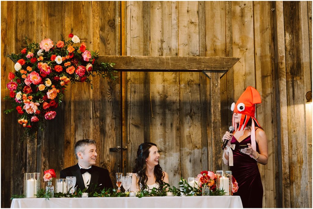 Snohomish wedding photographer gsquared weddings a person wearing a squid hat stands and speaks into a microphone at a wedding reception The couple sits at a decorated table smiling The backdrop is rustic wooden with a floral arrangement Serving seattle snohomish and washington state