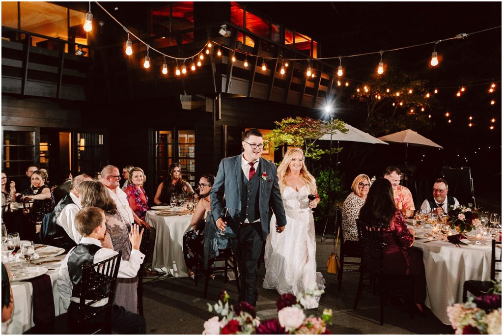 Snohomish wedding photographer gsquared weddings a bride and groom hold hands smiling as they walk through an outdoor wedding reception Guests are seated at round tables under string lights The evening setting includes a large building with warm lighting in the background Serving seattle snohomish and washington state
