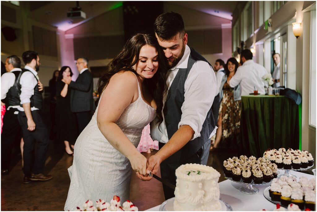 Snohomish wedding photographer gsquared weddings a couple wearing wedding attire is smiling and cutting a small cake together at a reception Around them other guests are mingling and tables are decorated with cupcakes and flowers The room is softly lit creating a joyful atmosphere Serving seattle snohomish and washington state