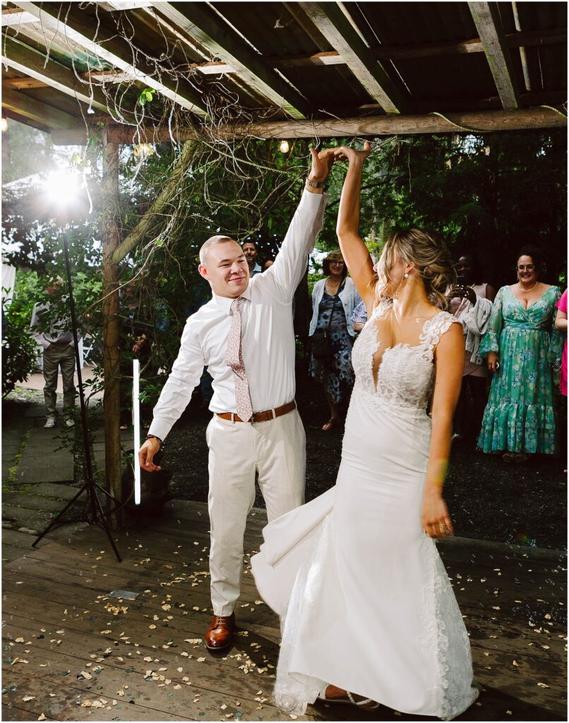 Snohomish wedding photographer gsquared weddings a couple dances under a wooden pergola The groom in a white shirt and tie twirls the bride who wears a white lace wedding dress Guests watch in the background The setting appears to be outdoors with greenery around Serving seattle snohomish and washington state