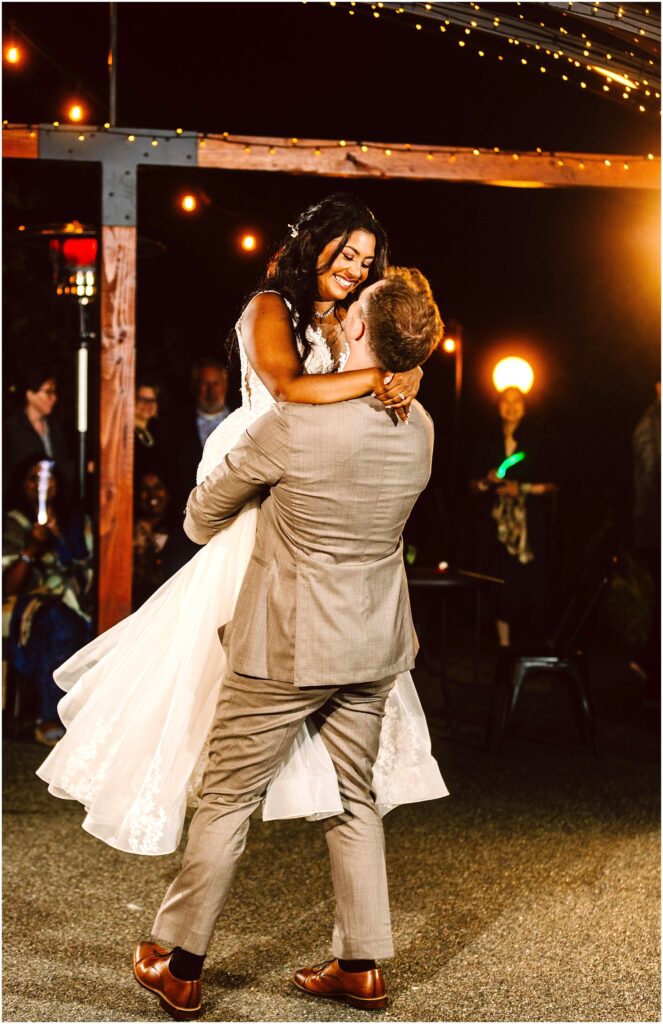 Snohomish wedding photographer gsquared weddings a groom in a light gray suit lifts the smiling bride in a white dress during their outdoor wedding dance They are surrounded by guests under warm glowing string lights at night Serving seattle snohomish and washington state