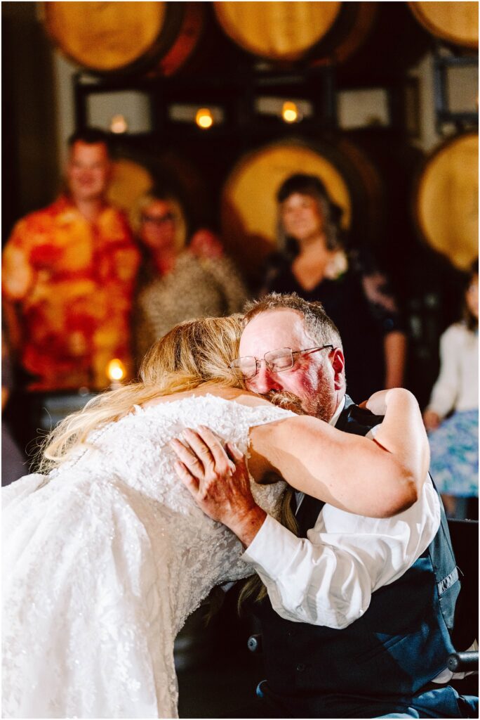 Snohomish wedding photographer gsquared weddings a bride in a white lace dress is hugging an older man wearing glasses and a vest They are in a warmly lit room with large barrels in the background Several people are softly focused in the background Serving seattle snohomish and washington state