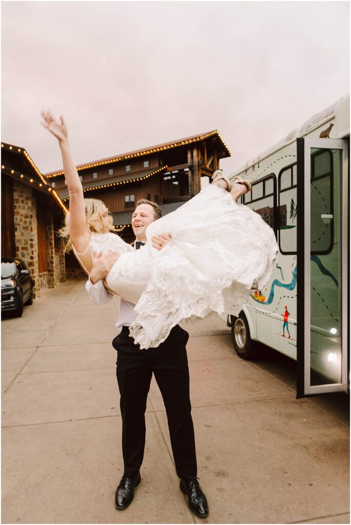 Snohomish wedding photographer gsquared weddings a man in a black suit lifts a woman in a white wedding dress joyfully in front of a decorated bus They are both smiling and the woman has one arm raised The setting appears to be outside near buildings with string lights Serving seattle snohomish and washington state