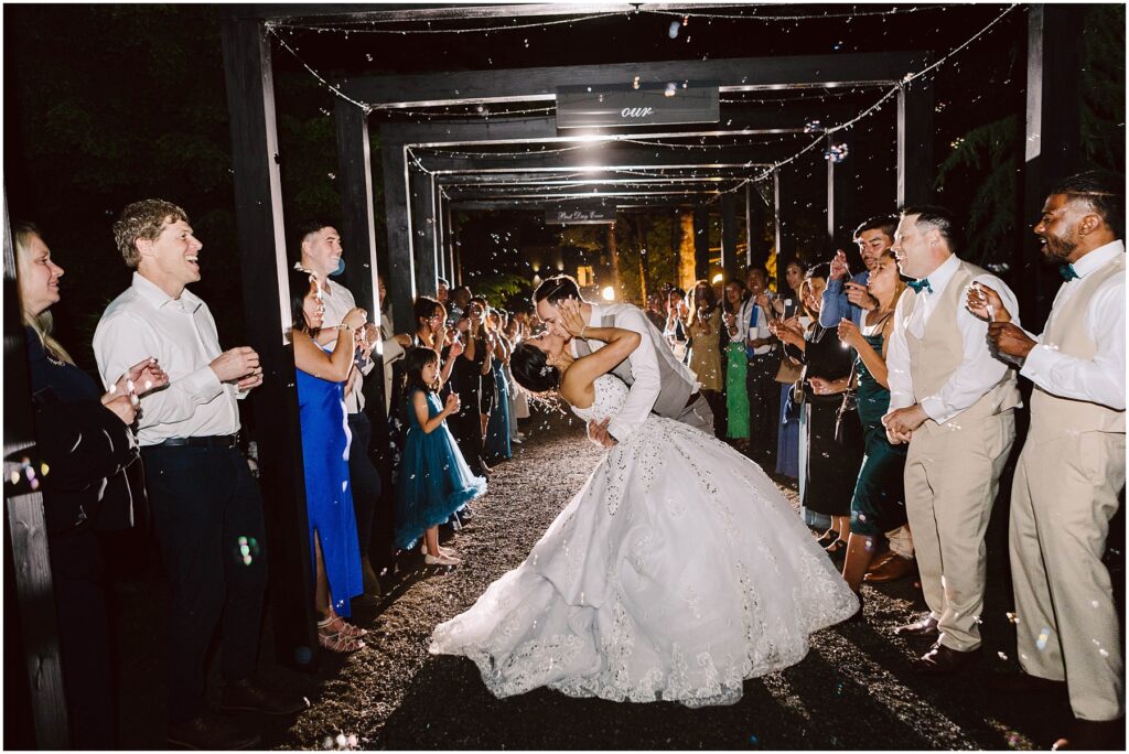 Snohomish wedding photographer gsquared weddings a bride and groom share a kiss under a decorated archway at night surrounded by applauding guests The bride wears a white gown and the groom is in a suit Bubbles fill the air adding a magical atmosphere to the scene Serving seattle snohomish and washington state