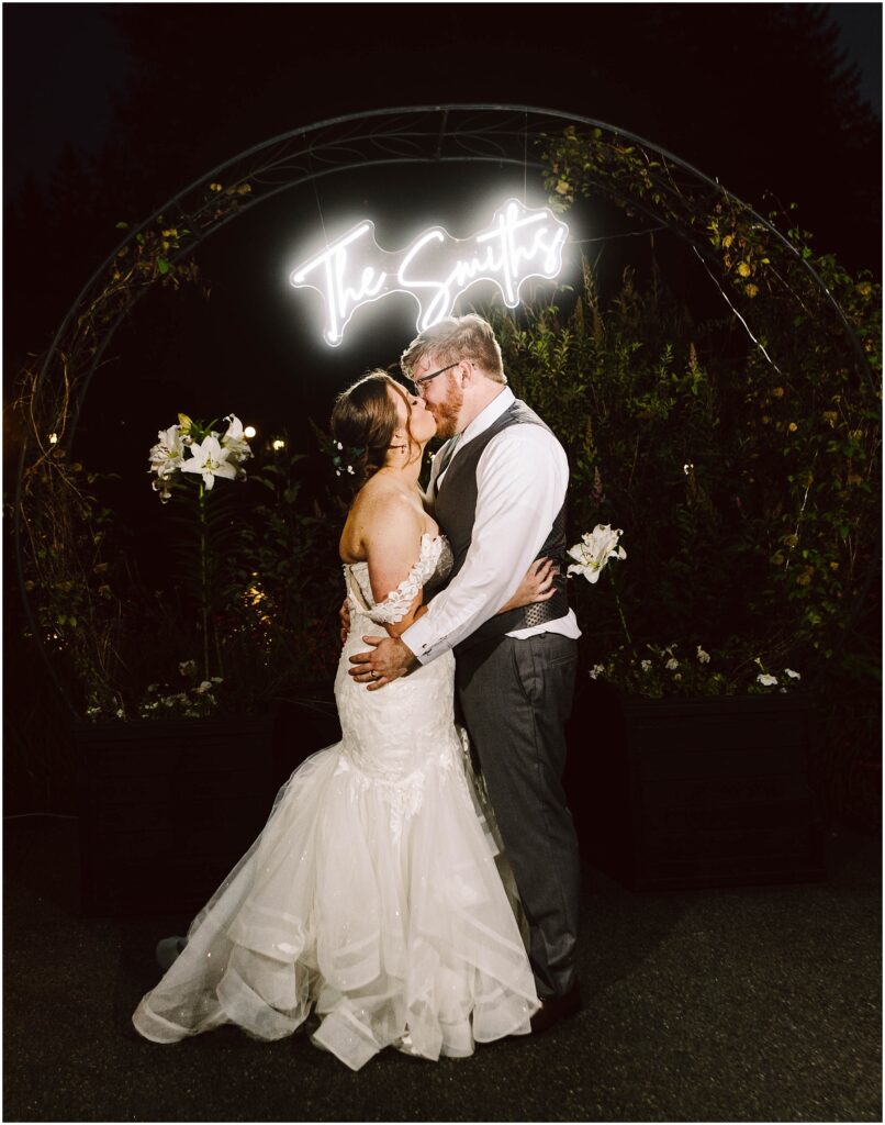 Snohomish wedding photographer gsquared weddings a bride and groom share a kiss under a neon sign reading the smiths The bride wears a white gown with lace details and the groom is in a vest and tie They are framed by an arch decorated with white flowers and greenery Serving seattle snohomish and washington state