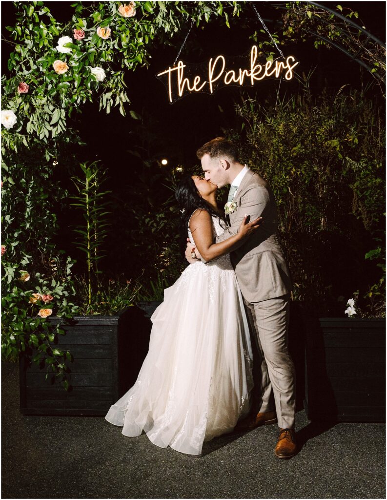 Snohomish wedding photographer gsquared weddings a bride and groom share a kiss under an arch adorned with flowers and greenery Above them is a neon sign displaying the parkers The bride wears a flowing white gown and the groom is in a light gray suit It is nighttime Serving seattle snohomish and washington state